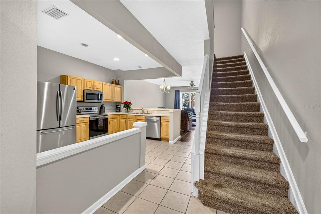 A kitchen with light wood cabinets and stainless steel appliances sits beside a carpeted staircase — 26624 CHIMNEY SPIRE LANE, WESLEY CHAPEL, FL