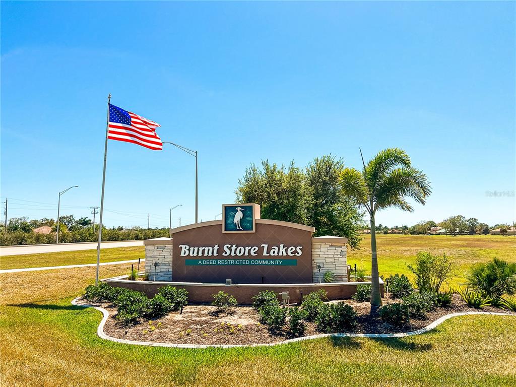A sign for Burnt Store Lakes stands in a grassy field with an American flag — 17283 CARROWAY LANE, PUNTA GORDA, FL