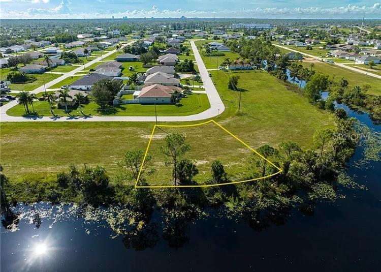 An aerial view shows a vacant, trapezoidal waterfront lot outlined in yellow near residential homes — 2117 NE 43RD LANE, CAPE CORAL, FL