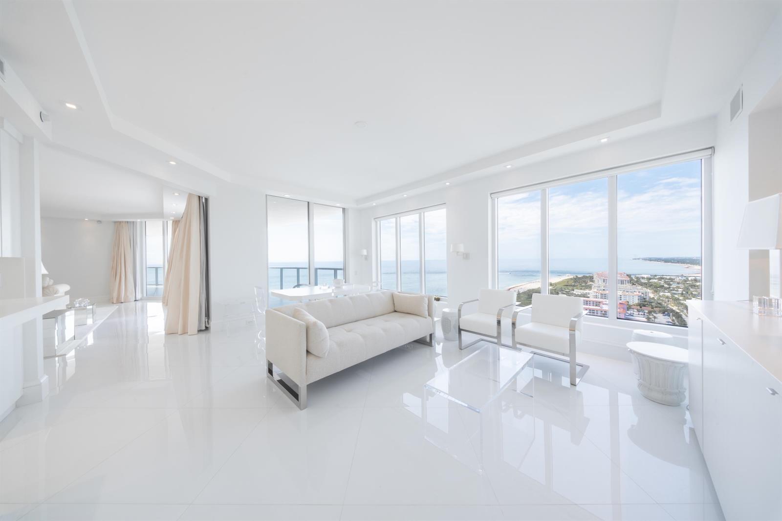 A bright, minimalist living room with white tile floors, modern furniture, and floor-to-ceiling ocean views — 2700 N Ocean Drive # 2503a, Riviera Beach FL 33404, RIVIERA BEACH, FL