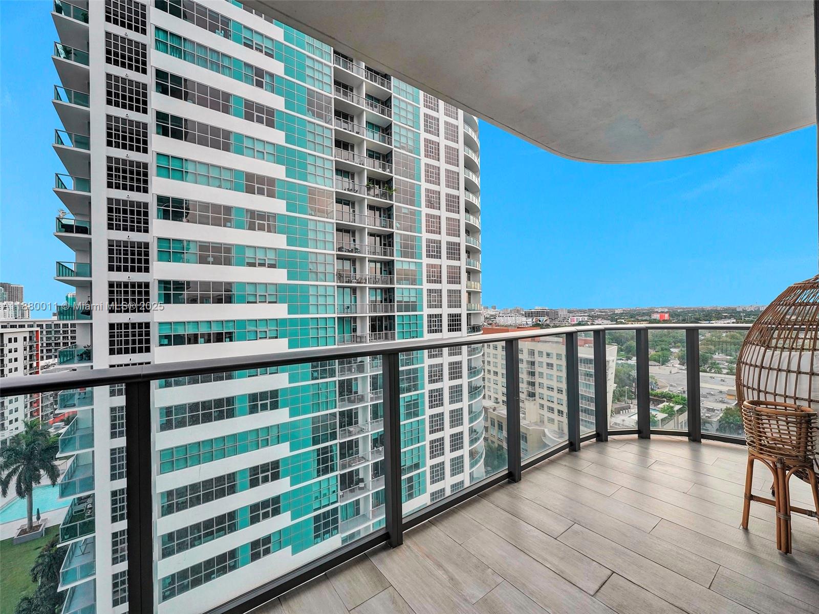 A balcony with wood-look tile flooring, a glass railing, and a wicker chair overlooks city buildings — 121 NE 34th St # 1601, Miami FL 33137, MIAMI, FL