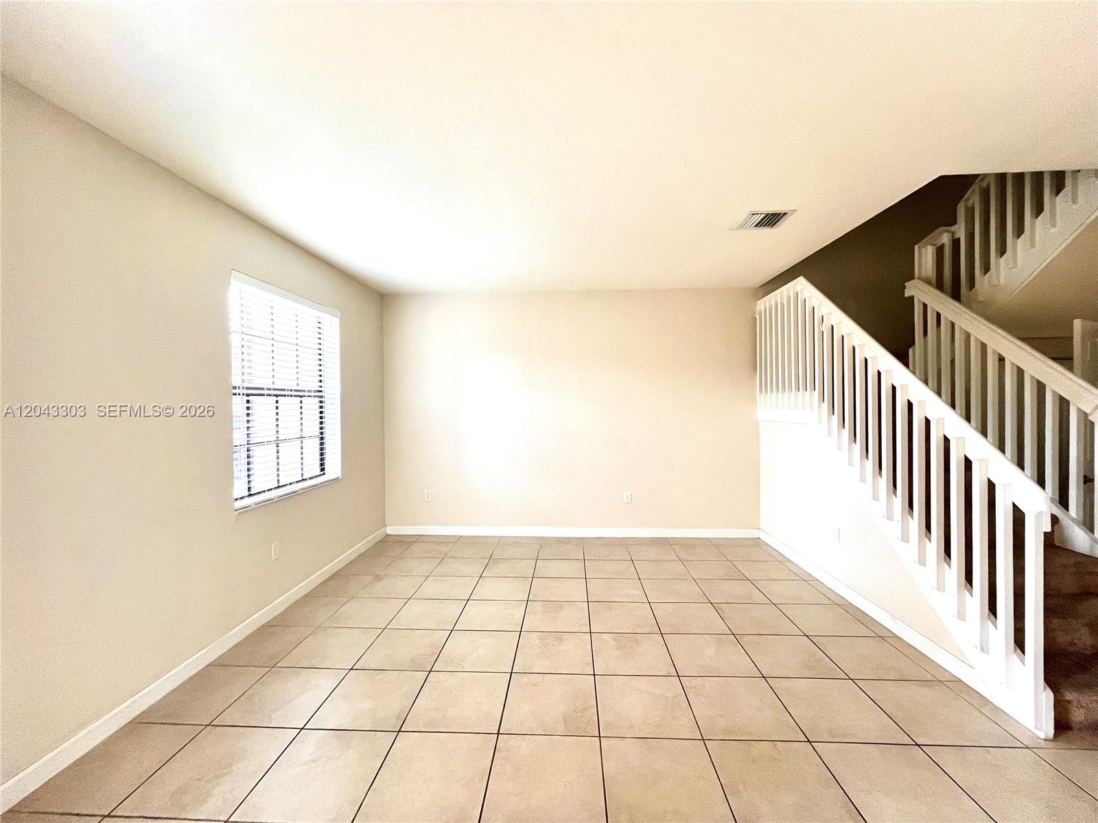 An empty room features beige tile flooring, light-colored walls, a window, and a wooden staircase — 9271 W 33rd Ln # 9271, Hialeah FL 33018, HIALEAH, FL