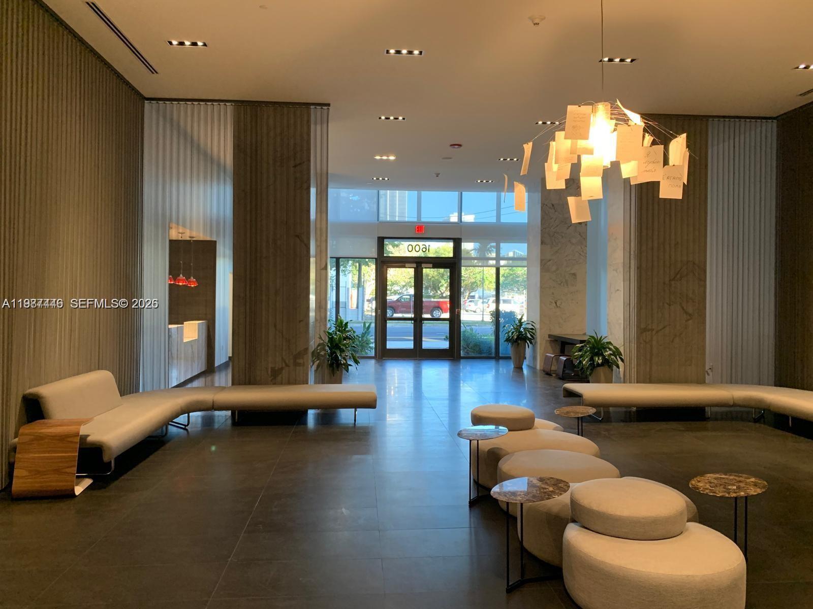 A modern building lobby features curved seating, circular ottomans, a paper-themed chandelier, and glass entrance doors — 1600 NE 1st Ave # 1715, Miami FL 33132, MIAMI, FL