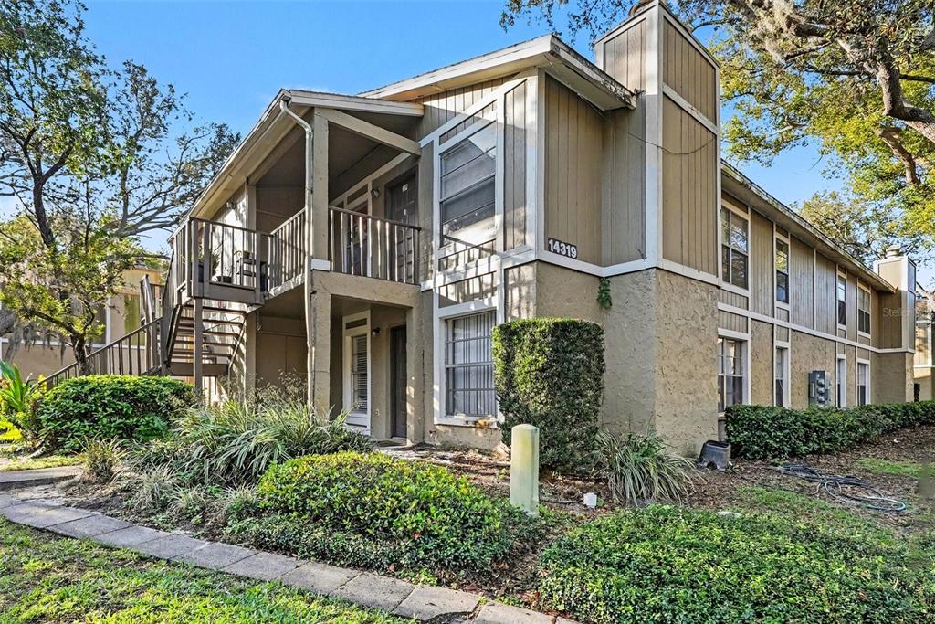 A two-story apartment building exterior features tan siding, a wooden staircase, and landscaped grounds — 14319 HANGING MOSS CIRCLE UNIT 101, TAMPA, FL