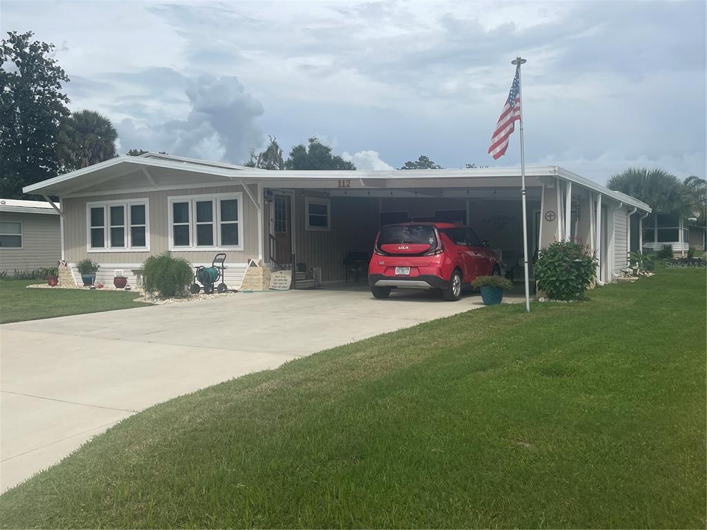 A single-story manufactured home with a carport, a red car, and an American flag — 112 LYONIA LANE, WILDWOOD, FL