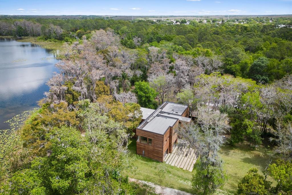 An aerial view shows a modern wooden house nestled in a wooded area beside a lake — 3807 S SEMORAN BOULEVARD, ORLANDO, FL