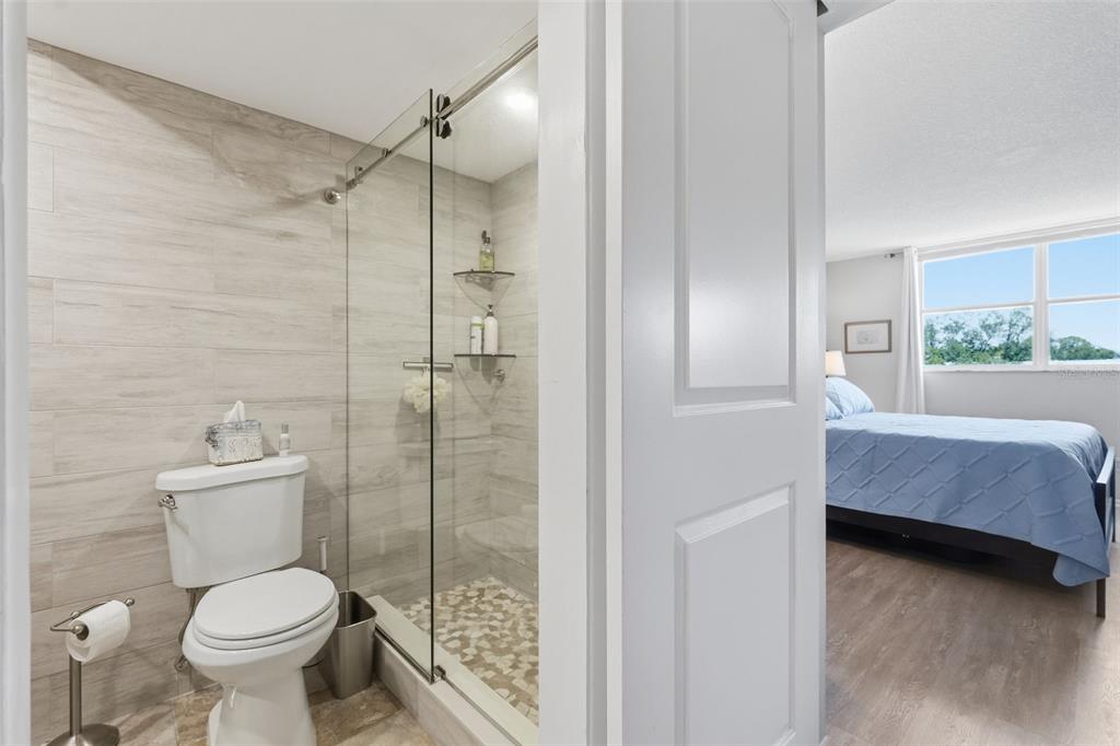 A bathroom with a glass-enclosed shower and toilet, adjacent to a bedroom with a bed — 4915 BAY STREET NE UNIT 323, ST PETERSBURG, FL