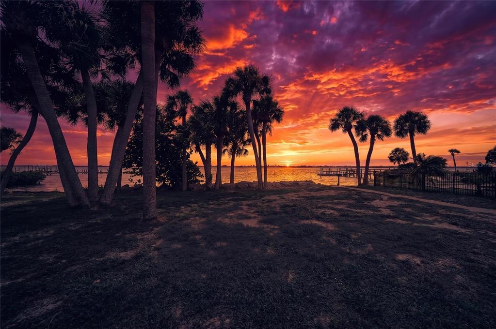 A vibrant sunset illuminates palm trees and a waterfront view from a grassy yard — 211 BAHIA VISTA DRIVE, ENGLEWOOD, FL