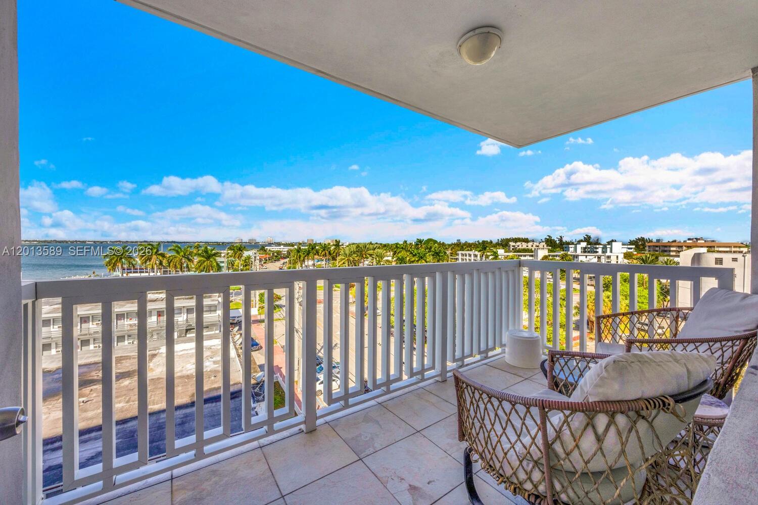 A balcony with white railings, tiled flooring, and wicker chairs overlooks a coastal city view — 2016 Bay Dr # 604, Miami Beach FL 33141, MIAMI BEACH, FL
