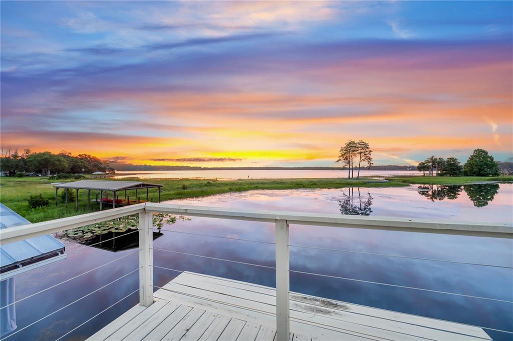 A wooden deck with a railing overlooks a lake at sunset with a boat dock — 4931 DORA DRIVE, MOUNT DORA, FL