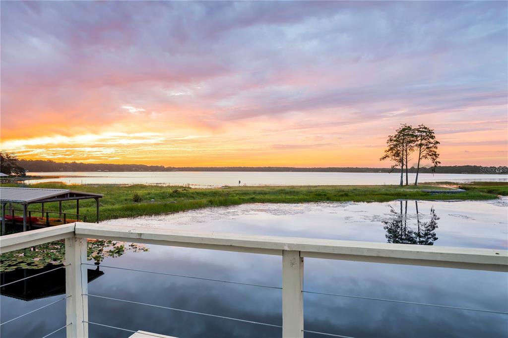 A wooden deck railing overlooks a lake at sunset with a boat dock nearby — 4931 DORA DRIVE, MOUNT DORA, FL