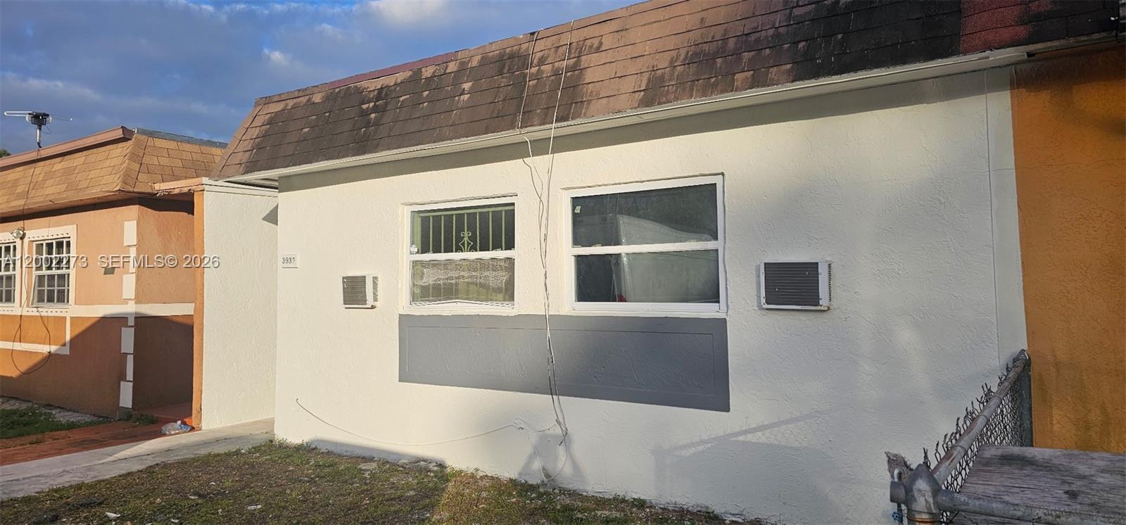 The exterior of a single-story house features white stucco walls, two windows, and two wall-mounted air conditioners — 3932 NW 207th St Rd, Miami Gardens FL 33055, MIAMI GARDENS, FL