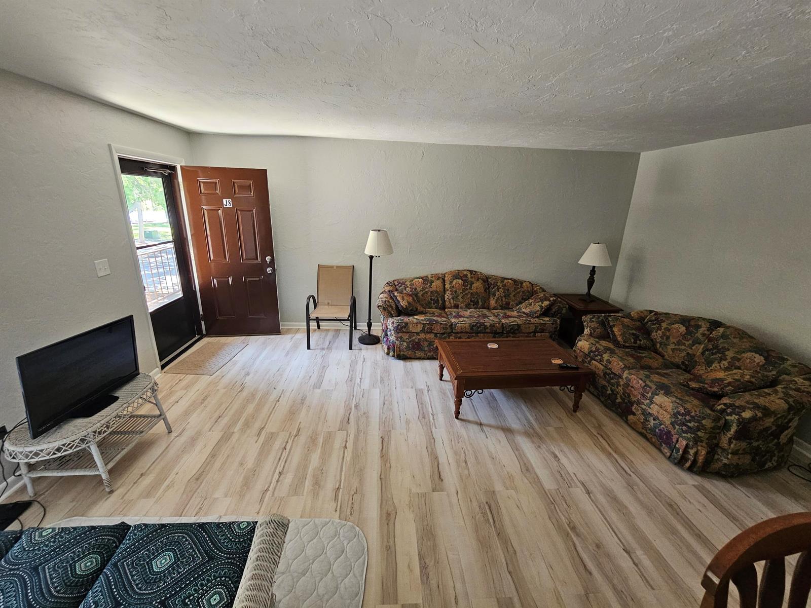 A living room features light wood-look flooring, two floral sofas, and a brown entry door — 3124 SE Us Highway 441 # J8, Okeechobee FL 34974, OKEECHOBEE, FL
