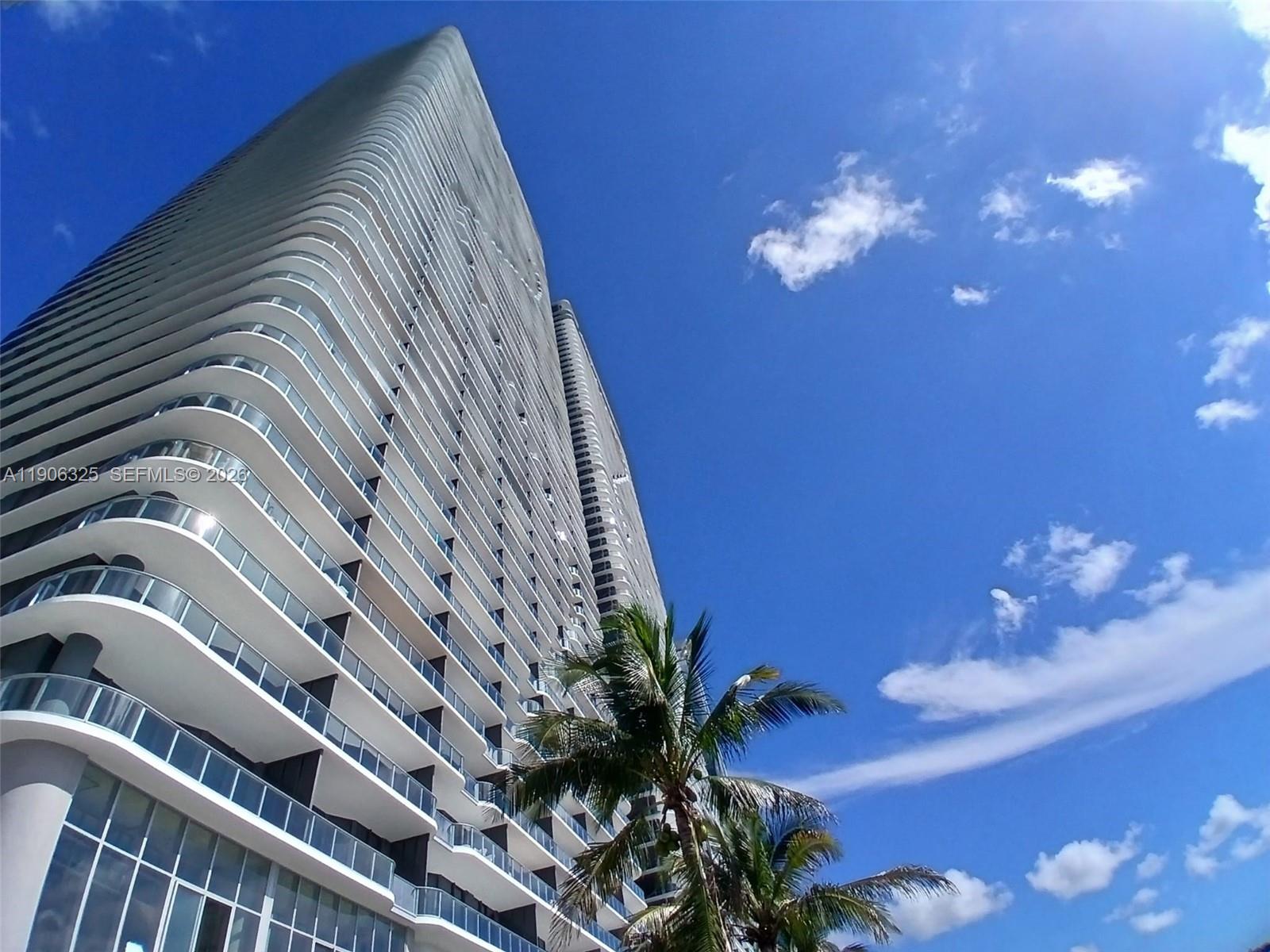A low-angle view of a tall, modern residential skyscraper with balconies against a blue sky — 700 NE 24 # 203, Miami FL 33137, MIAMI, FL