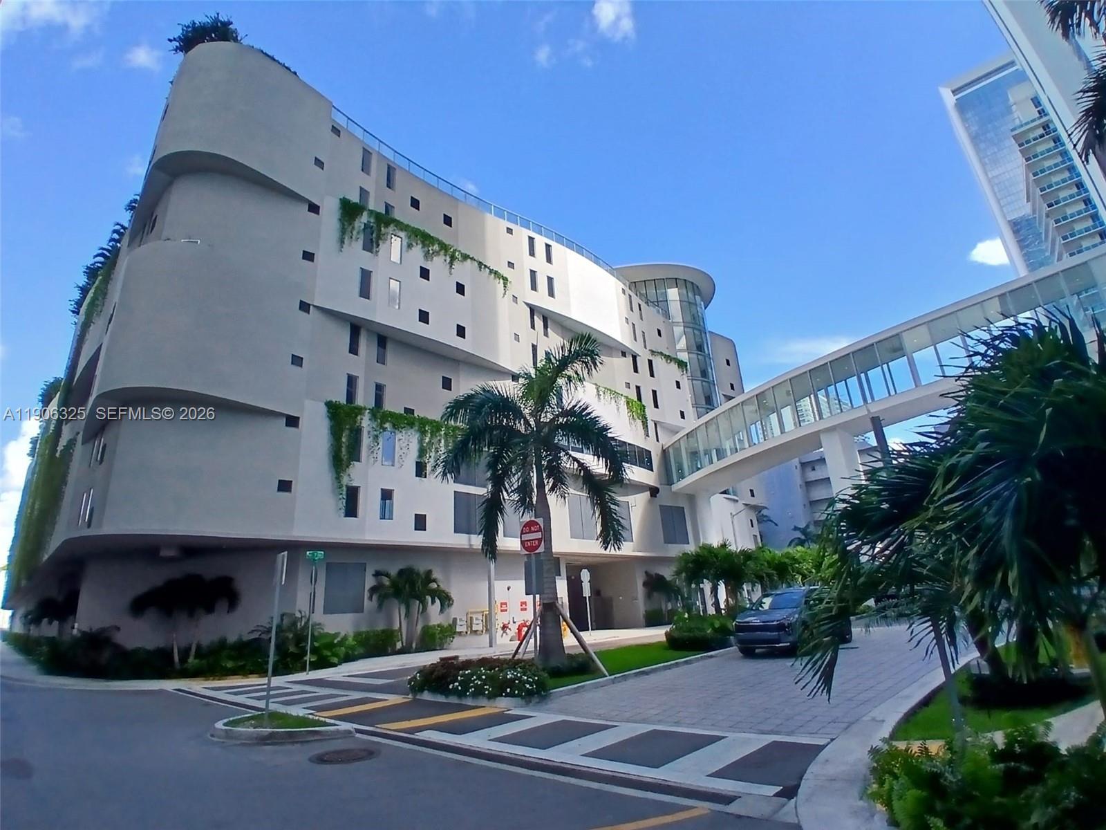 A modern multi-story building with a glass skybridge and a palm tree in the foreground — 700 NE 24 # 203, Miami FL 33137, MIAMI, FL