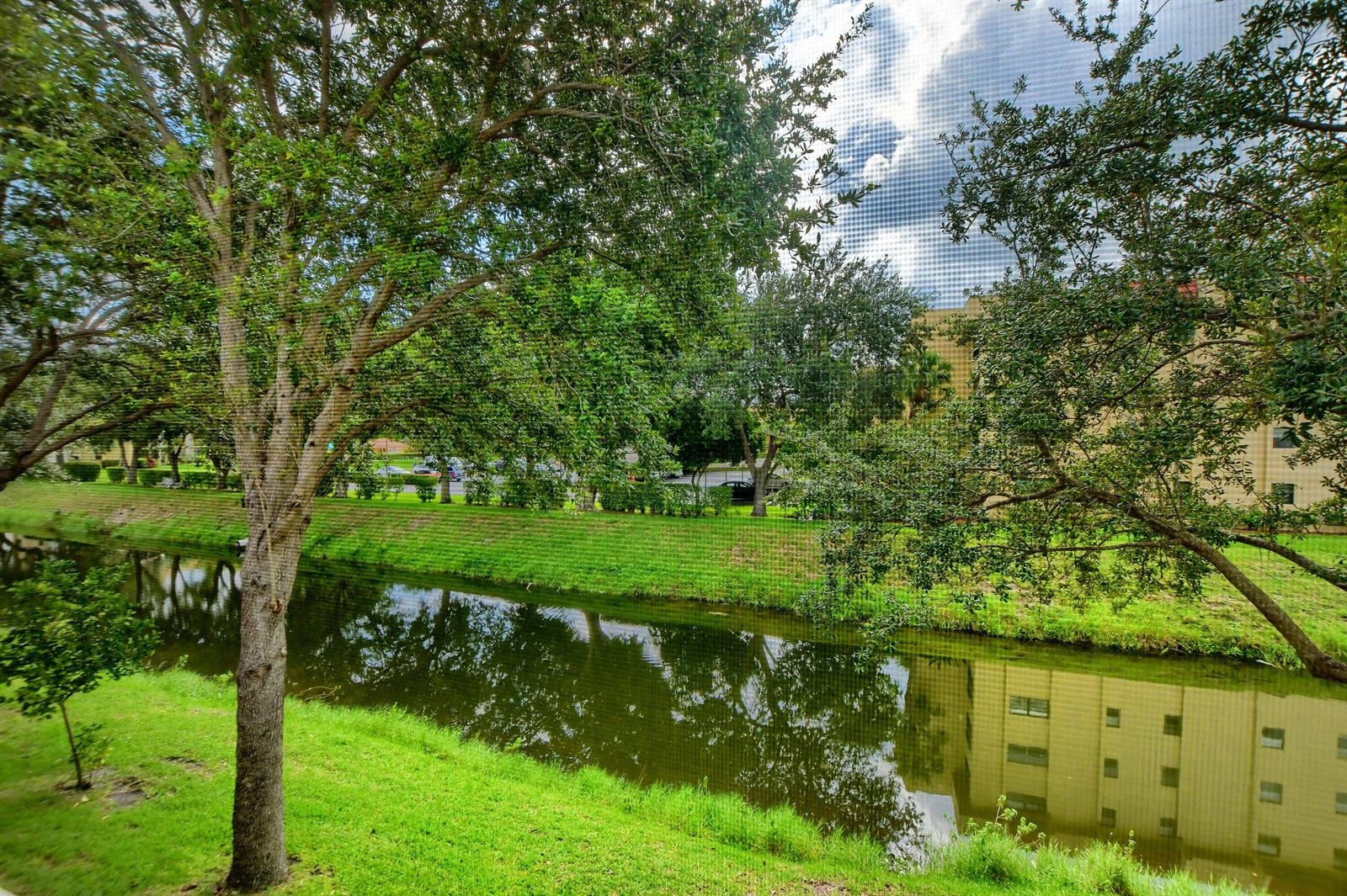 A view of a grassy canal bank with trees and a building reflection in the water — 9826 Marina Boulevard # 1027, Boca Raton FL 33428, BOCA RATON, FL