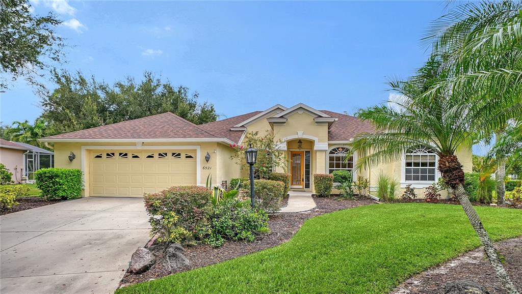 A single-story suburban house with a two-car garage, tan stucco exterior, and a palm tree — 6521 SUNDEW COURT, BRADENTON, FL