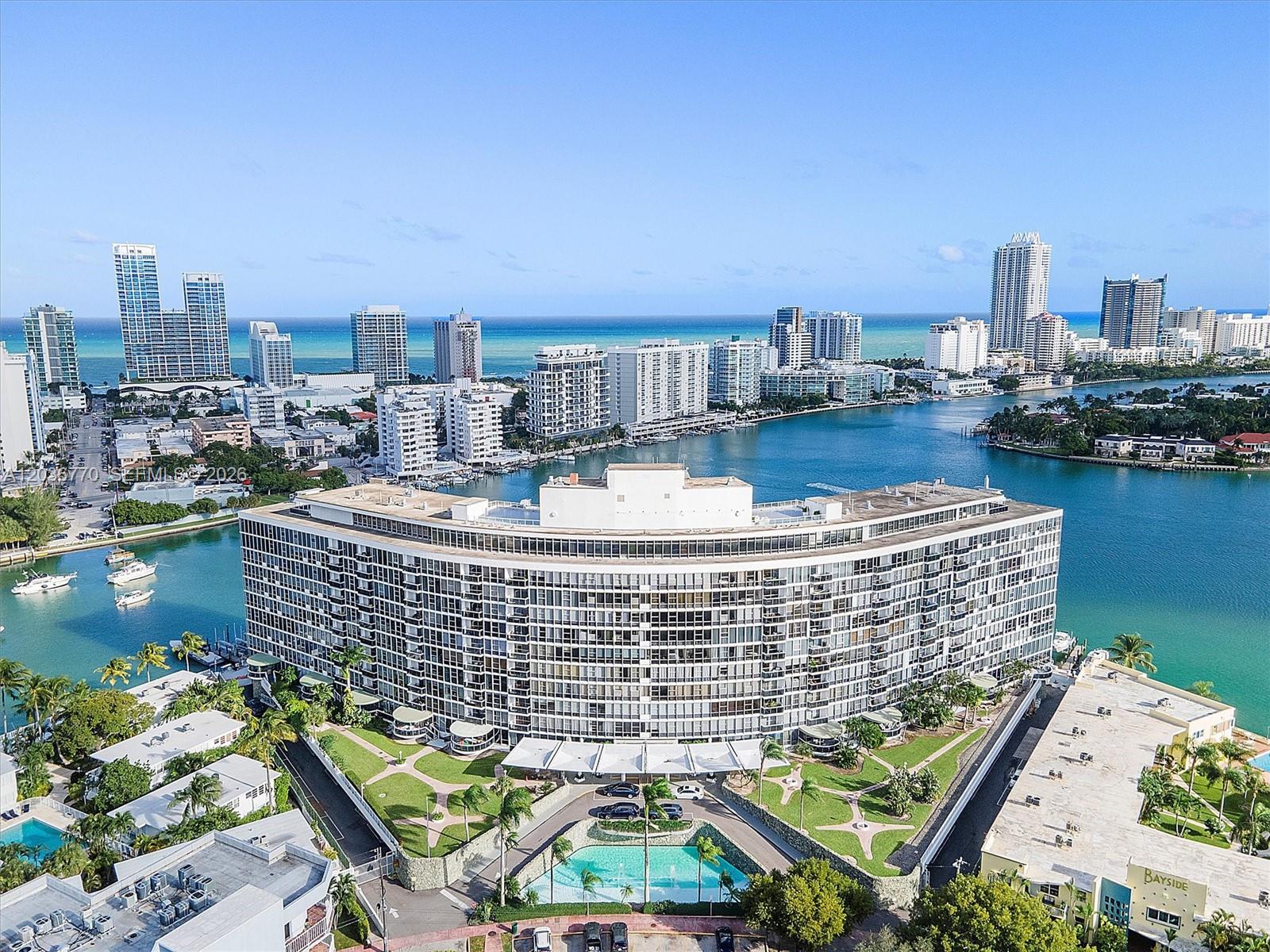 An aerial view shows a large, curved residential building surrounded by water and city skyline — 900 Bay Dr # 215, Miami Beach FL 33141, MIAMI BEACH, FL