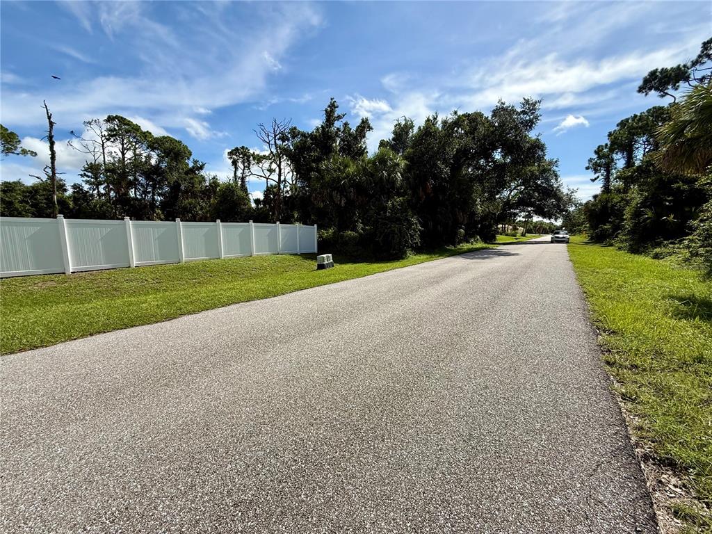 A paved road leads past a white vinyl fence and dense trees under a blue sky — 16046 GALENA AVE, PORT CHARLOTTE, FL