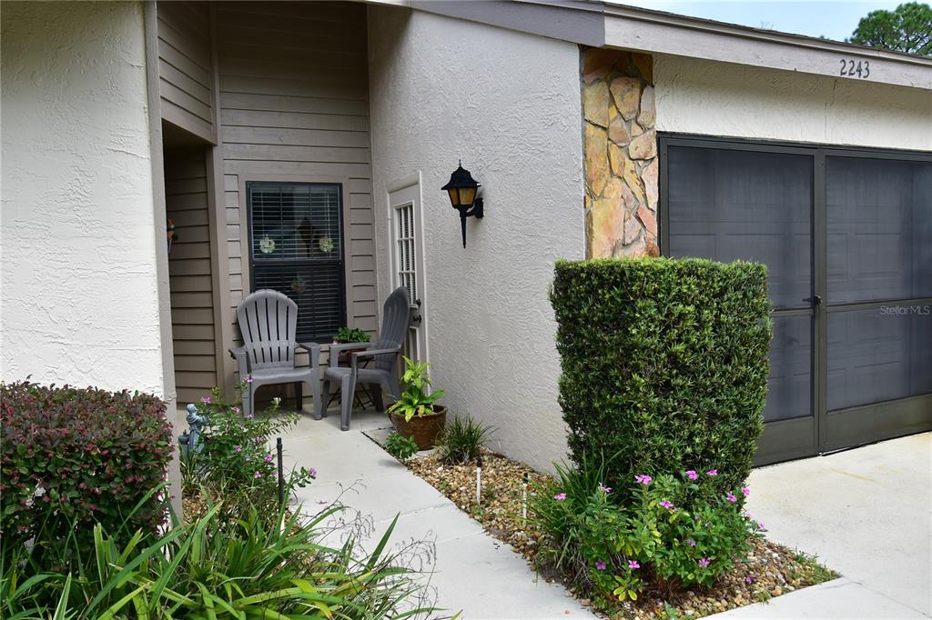 A concrete walkway leads to a covered entryway with two chairs and a garage door — 2243 WYNDAM DRIVE, SPRING HILL, FL