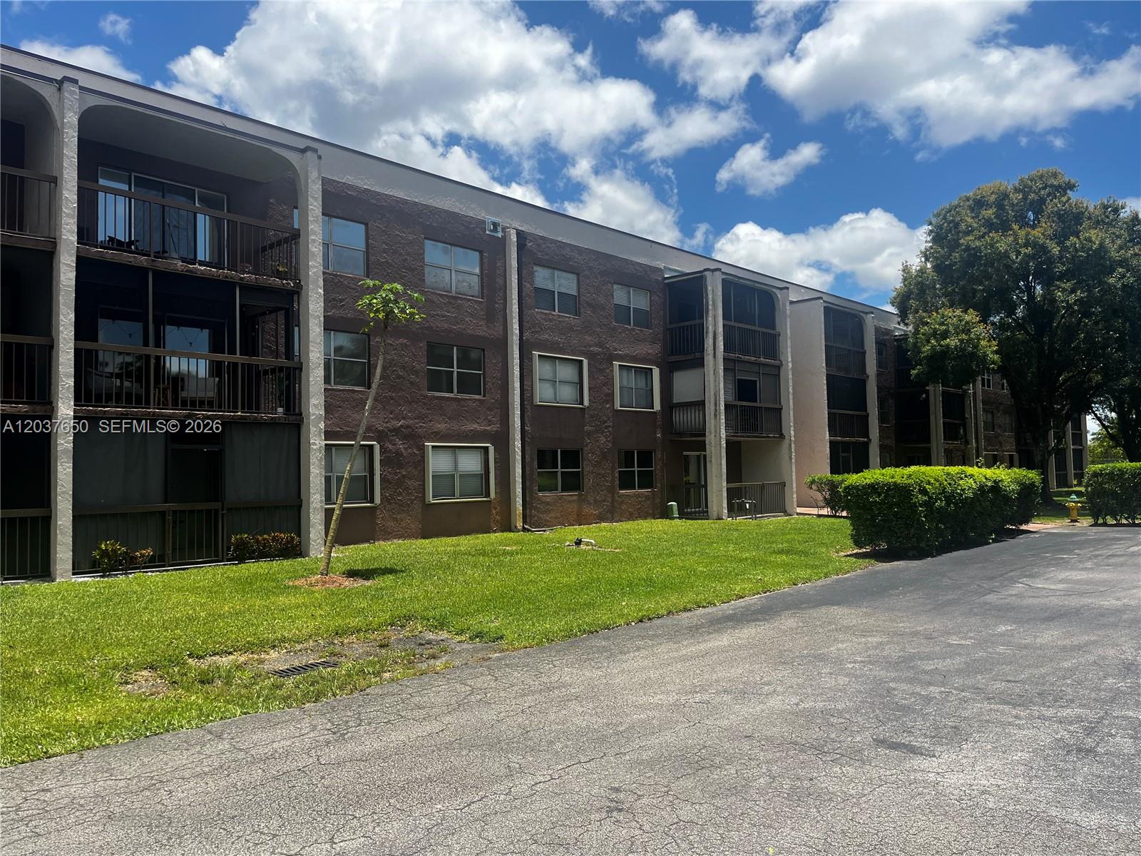 A three-story brown apartment building with balconies and a grassy lawn under a cloudy sky — 7000 Nova Dr # 103E, Davie FL 33317, DAVIE, FL