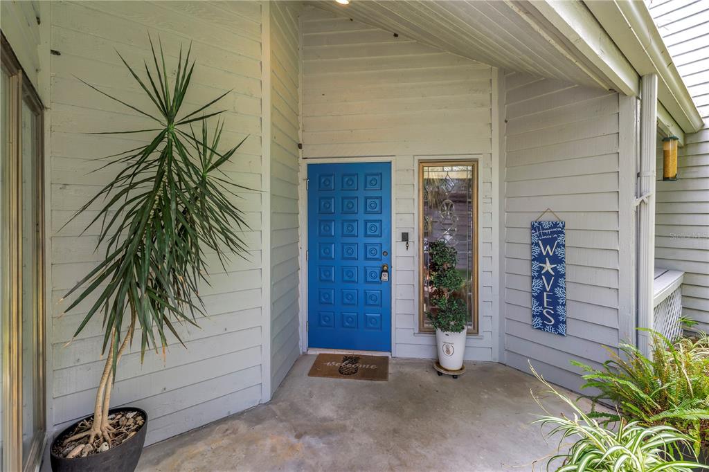 A blue front door with a sidelight window sits on a covered concrete porch — 10495 LONGWOOD DRIVE, SEMINOLE, FL