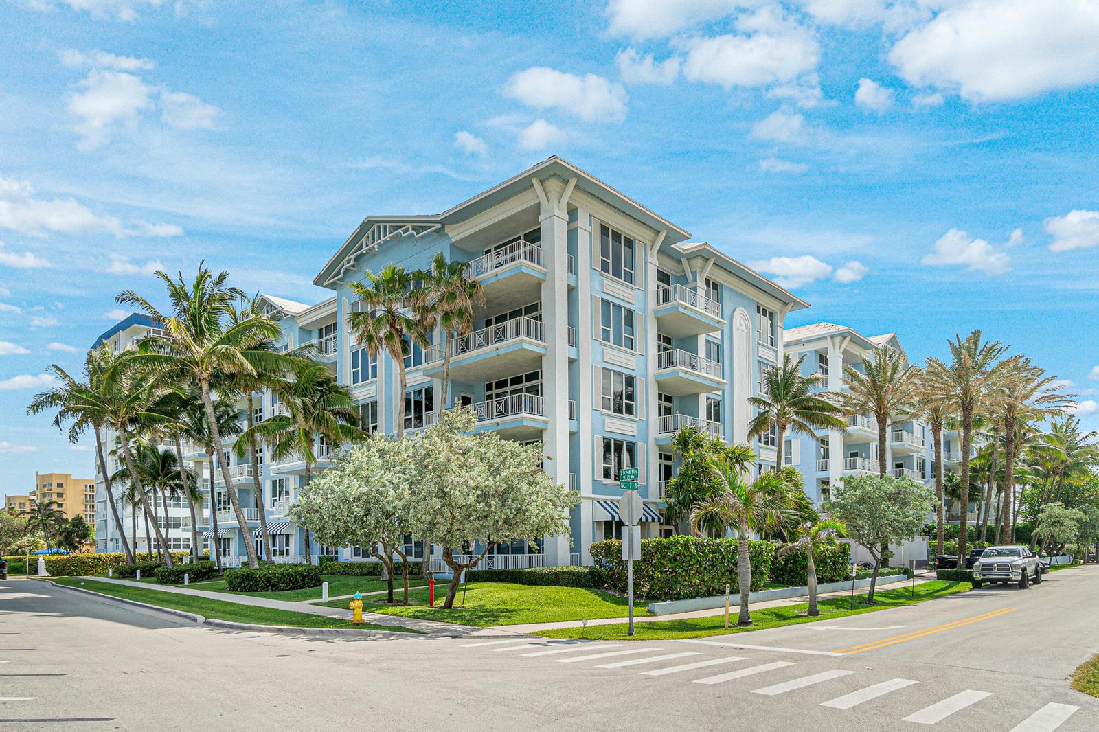 A multi-story blue apartment building with balconies stands on a corner lot with palm trees — 701 SE 21st Avenue # 301, Deerfield Beach FL 33441, DEERFIELD BEACH, FL