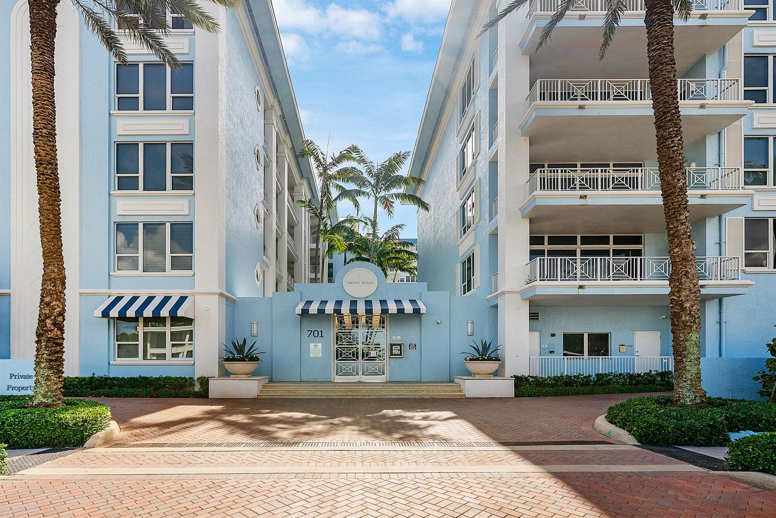 The exterior of a light blue multi-story apartment building with palm trees and a brick driveway — 701 SE 21st Avenue # 301, Deerfield Beach FL 33441, DEERFIELD BEACH, FL