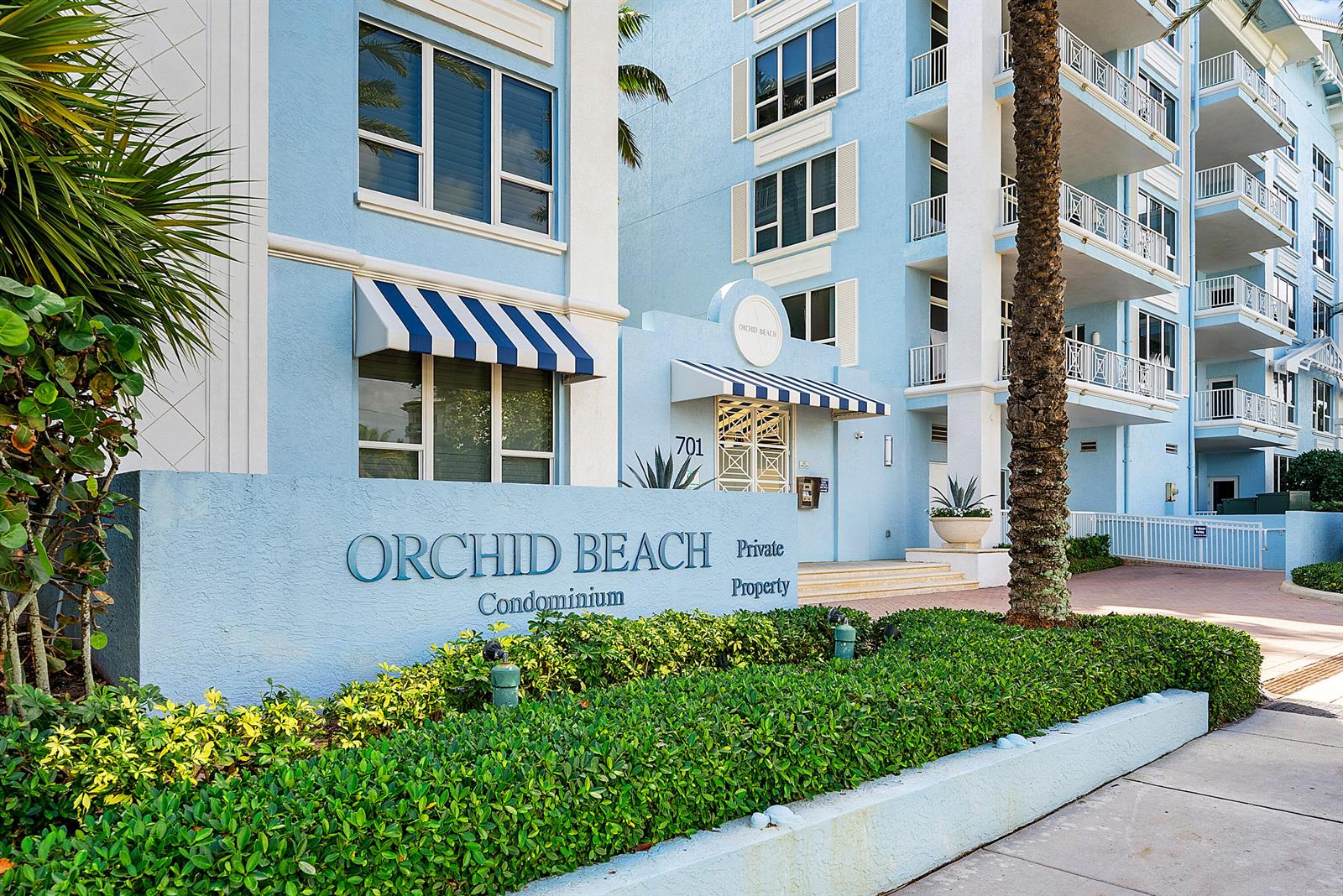 The exterior of a light blue condominium building features striped awnings and a landscaped sign — 701 SE 21st Avenue # 301, Deerfield Beach FL 33441, DEERFIELD BEACH, FL