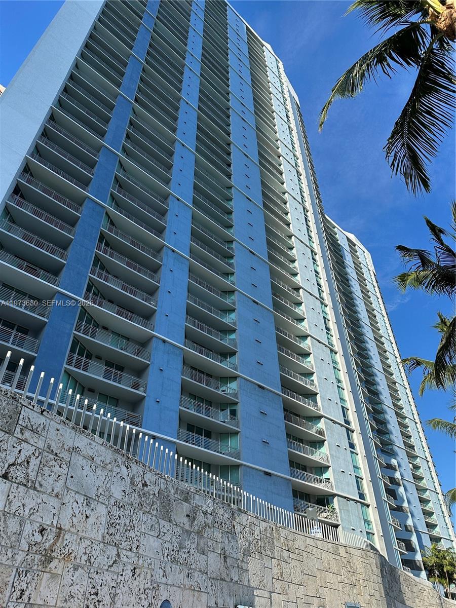 A low-angle view of a tall, blue residential high-rise building with numerous balconies — 335 S Biscayne Blvd # 1002, Miami FL 33131, MIAMI, FL
