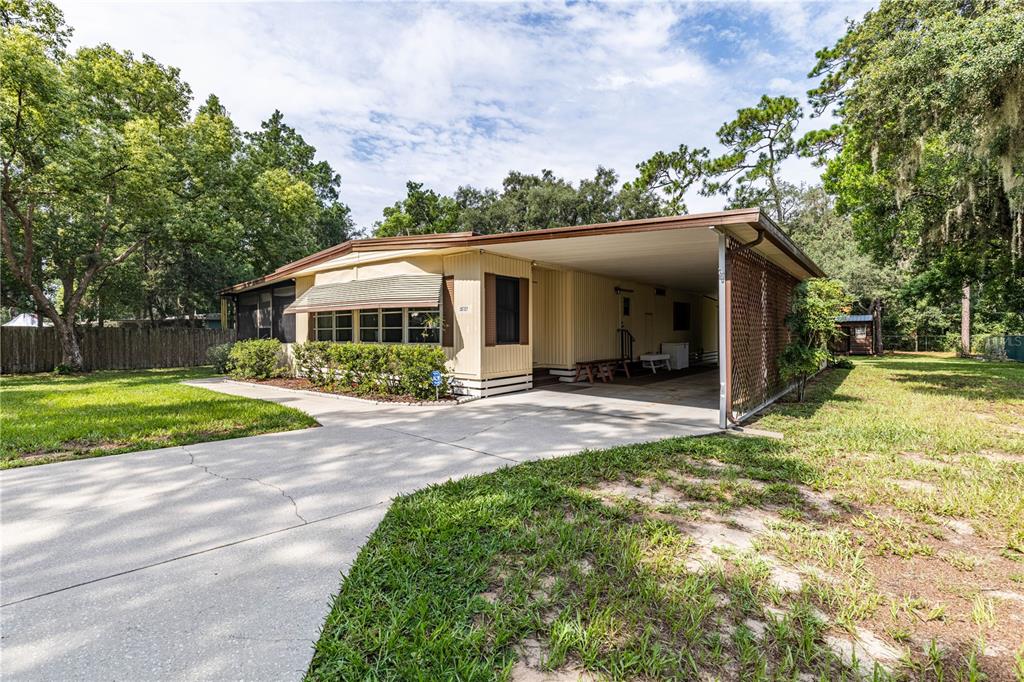 A single-story manufactured home with a concrete driveway, attached carport, and surrounding trees — 35737 SHELLEY DRIVE, LEESBURG, FL