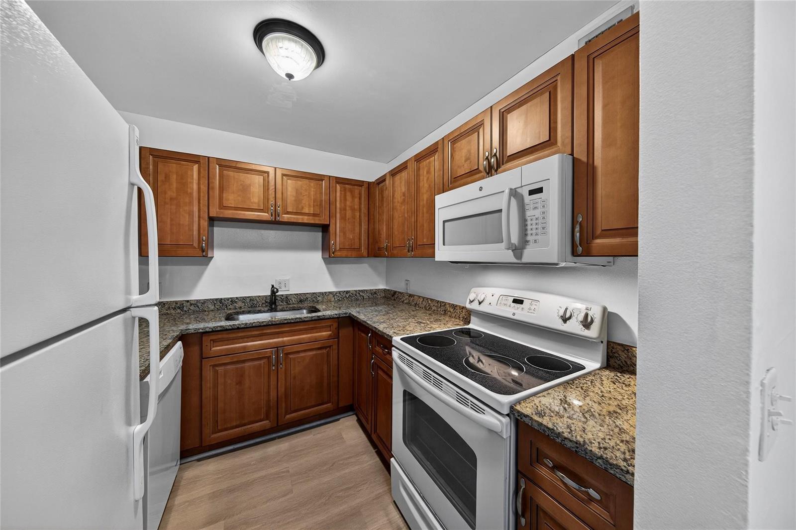A galley kitchen features wood cabinets, granite countertops, white appliances, and light wood-look flooring — 2201 NE 36th Street # 105, Lighthouse Point FL 33064, LIGHTHOUSE POINT, FL