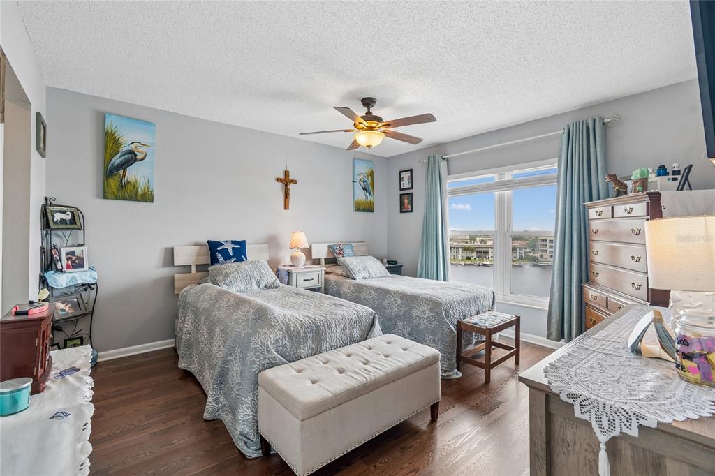 A bedroom features two twin beds, a tufted bench, a dresser, and a waterfront window view — 4939 FLORAMAR TERRACE UNIT 610, NEW PORT RICHEY, FL