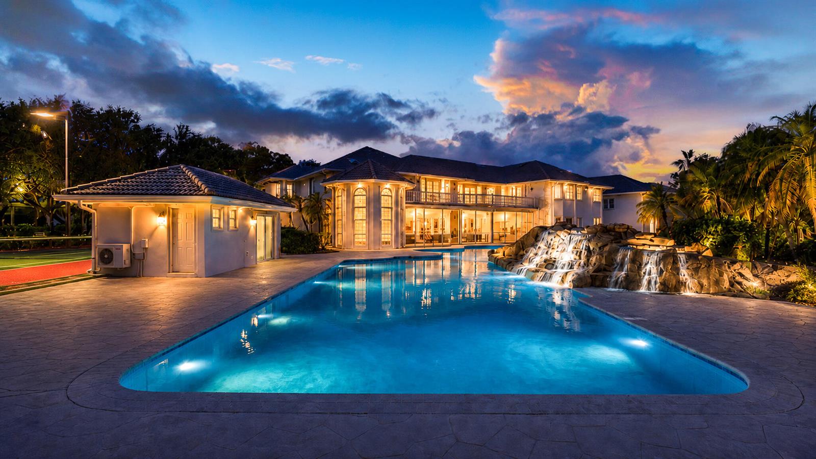 A large house with a swimming pool, waterfall, and tennis court at dusk — 1800 Eagle Trace Boulevard, Coral Springs FL 33071, CORAL SPRINGS, FL