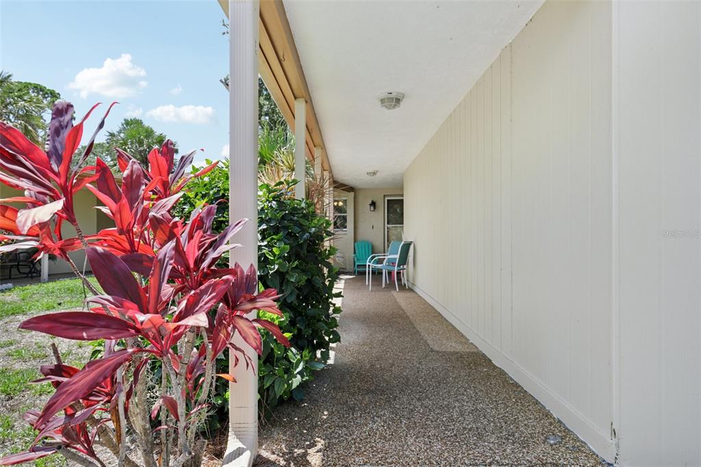 A covered outdoor walkway features a textured concrete floor, light-colored siding, and patio chairs — 7172 GREY SQUIRREL BOULEVARD UNIT 81, SARASOTA, FL