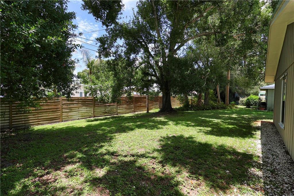 A grassy backyard features a large tree, a horizontal wood fence, and a house exterior — VERO BEACH, FL