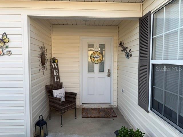 A covered front porch features a white door, a wicker chair, and decorative wall hangings — 7618 SE 171ST SUN VALLEY PLACE, THE VILLAGES, FL