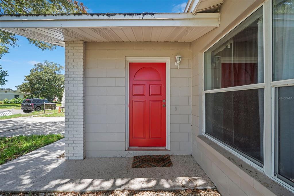 A red front door is centered on a beige concrete block wall under a porch — 1010 ILLINOIS ROAD, COCOA, FL