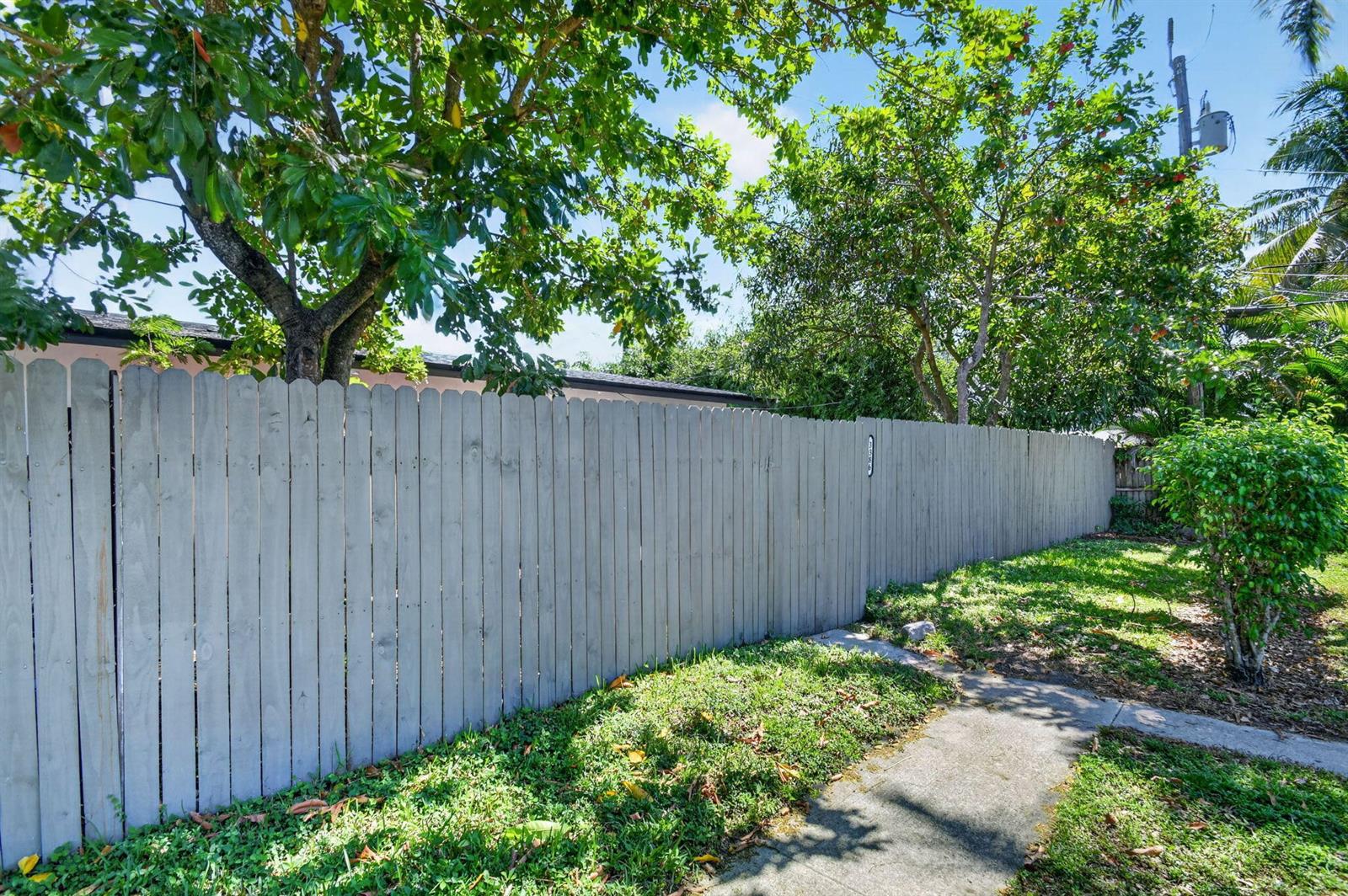A wooden privacy fence borders a grassy yard with a concrete walkway and mature trees — 3382 Rudolf Road, Lake Worth FL 33461, LAKE WORTH, FL