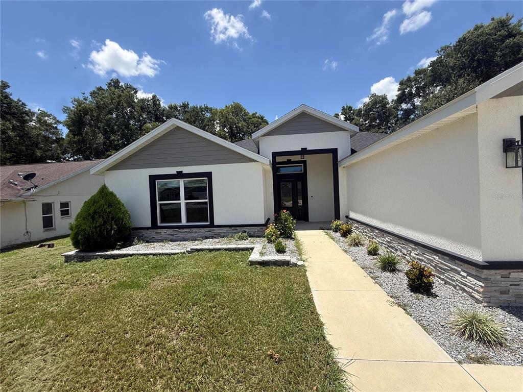 A modern single-story house exterior features white stucco, gray siding, and stone veneer accents — 16382 SE 83RD AVENUE, SUMMERFIELD, FL