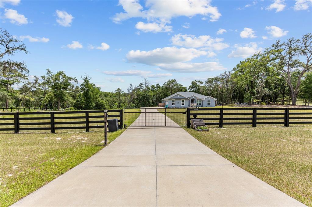 A concrete driveway leads through a black metal gate toward a house surrounded by trees — 10254 SE 118TH LANE, BELLEVIEW, FL