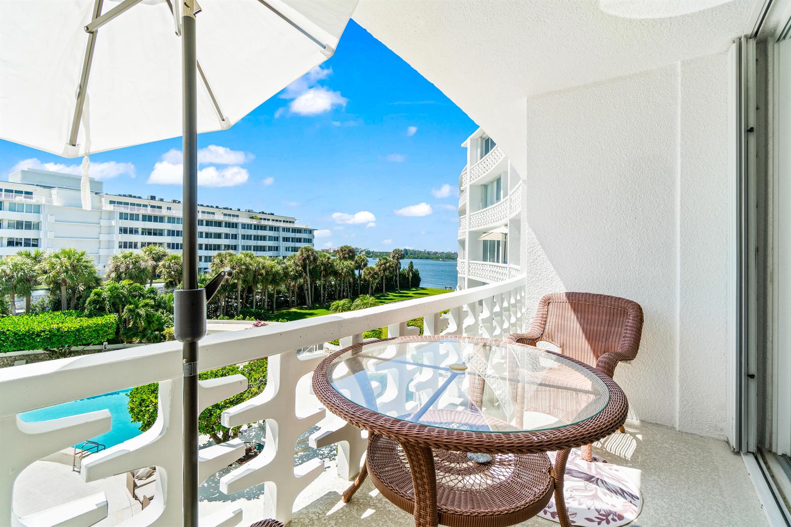 A balcony features a glass-topped wicker table, chairs, and an umbrella overlooking a pool and water — 2760 S Ocean Boulevard # 313, Palm Beach FL 33480, PALM BEACH, FL