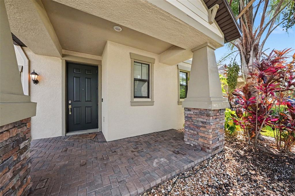 A covered front porch features stone-based columns, a dark door, and brick paver flooring — 10032 SAGE CREEK DRIVE, SUN CITY CENTER, FL