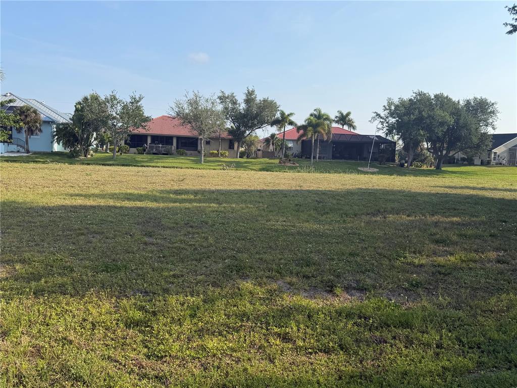A large, grassy vacant lot sits in front of several residential houses under a clear sky — 24336 SAN RAFAEL ROAD, PUNTA GORDA, FL