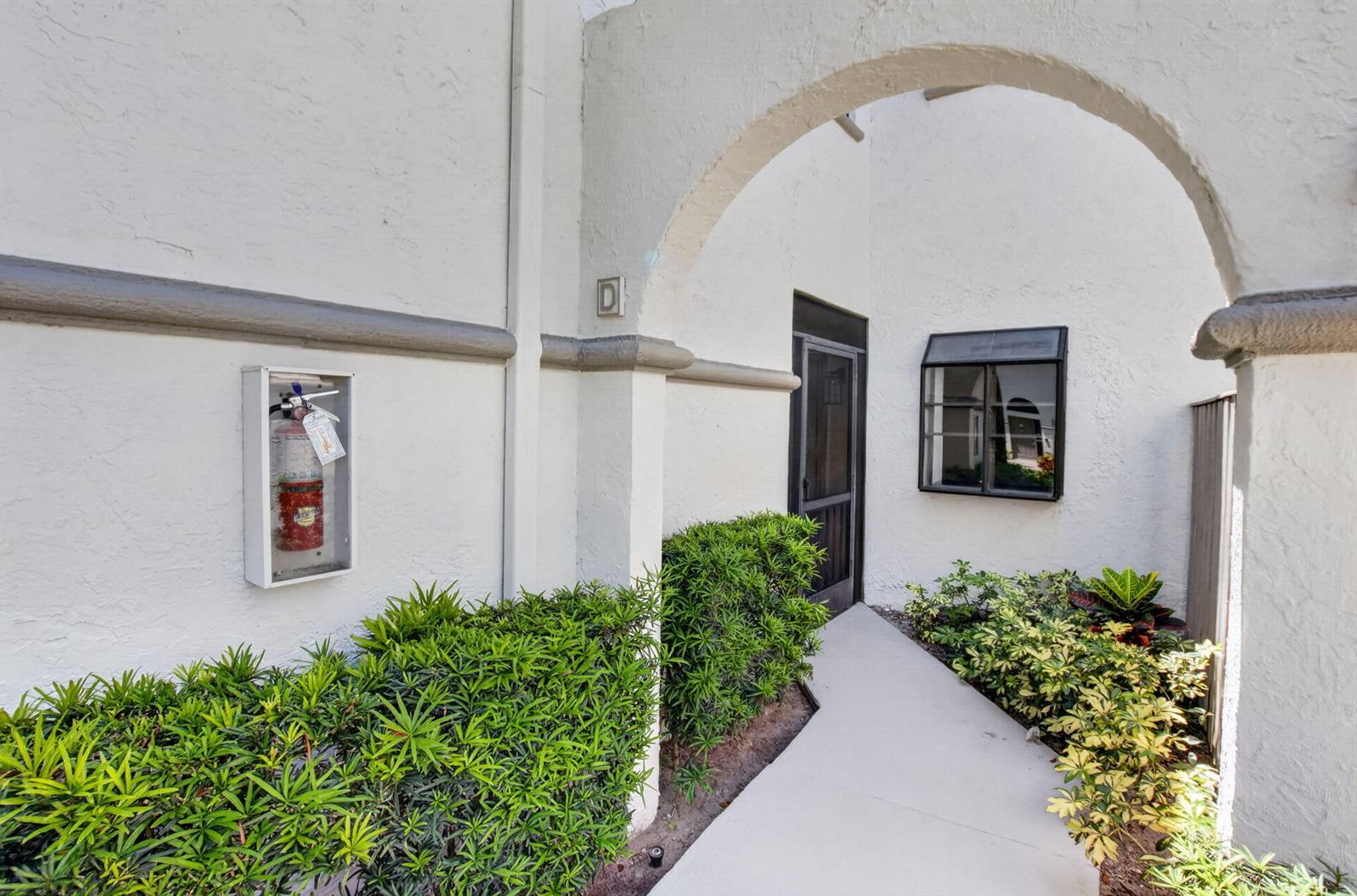 An exterior view of a building entrance with a fire extinguisher, landscaping, and an archway — 5259 Europa Drive # D, Boynton Beach FL 33437, BOYNTON BEACH, FL