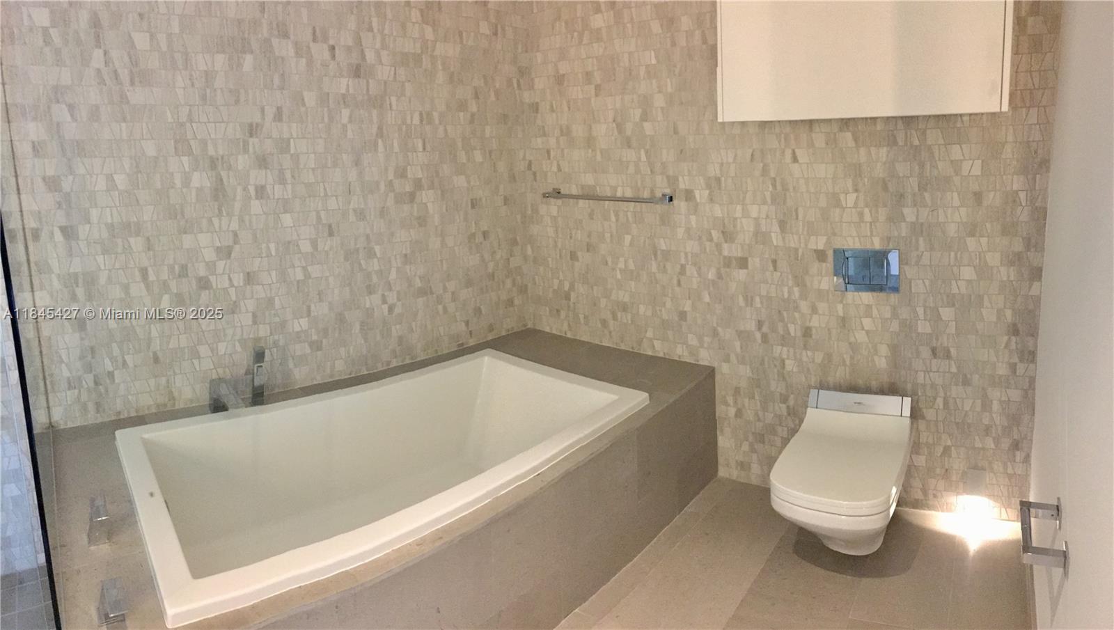 A modern bathroom features a built-in bathtub, a wall-mounted toilet, and textured stone wall tiles — 88 SW 7th St # 2305, Miami FL 33130, MIAMI, FL