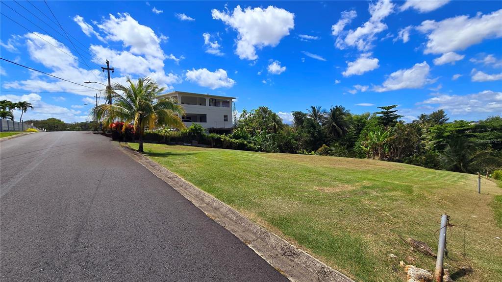 A paved road runs alongside a grassy, sloped lot with a multi-story house in the background — CALLE CEMI URB. HACIENDAS DEL CARIBE UNIT B6, TOA ALTA, PR