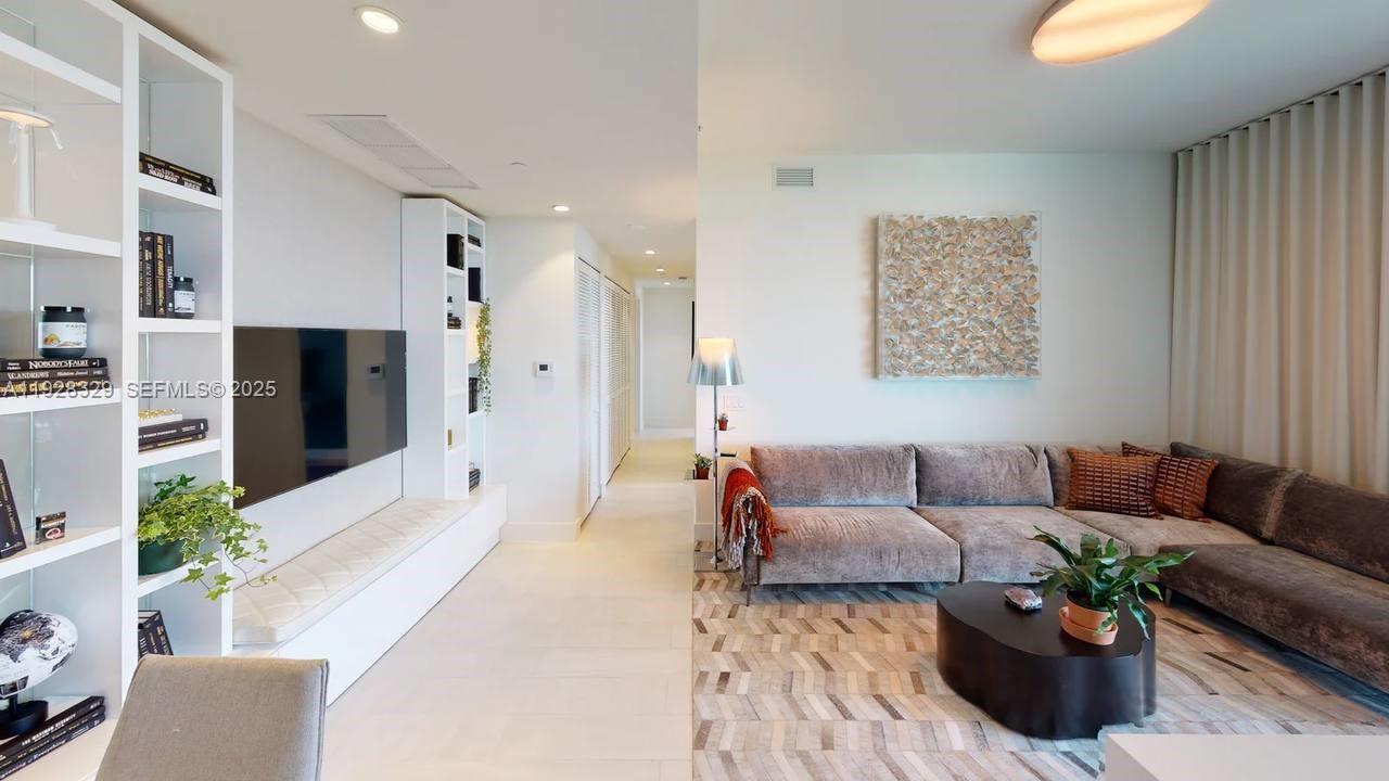 A modern living room features a large sectional sofa, a wall-mounted television, and built-in shelving — 2000 Metropica Way # 1701, Sunrise FL 33323, SUNRISE, FL