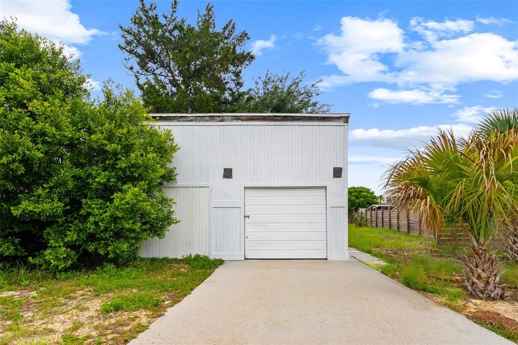 A white detached garage with a single roll-up door stands beside a large green tree — 21316 SUNSET AVENUE, PANAMA CITY BEACH, FL