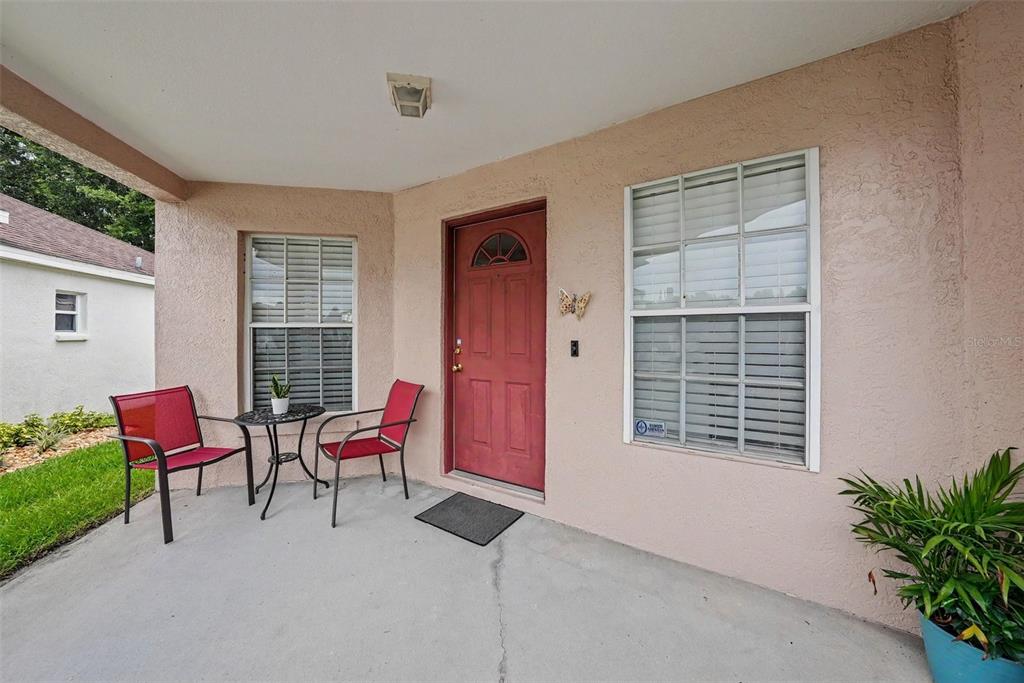 A covered concrete porch features a red front door, two red chairs, and a small table — 26351 WHIRLAWAY TERRACE, WESLEY CHAPEL, FL